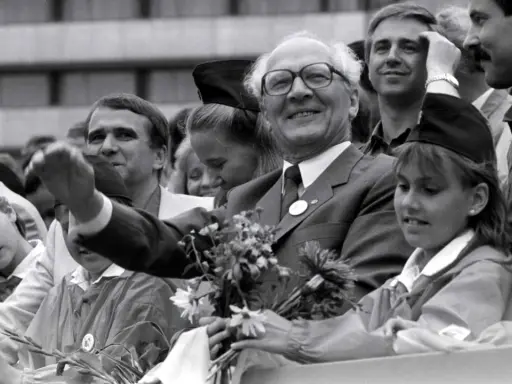 Ein Mann mit Brille winkt lächelnd, umgeben von Menschen. War die DDR eine Diktatur? Diese Szene zeigt eine öffentliche Veranstaltung, vielleicht ein Zeichen der Hoffnung.