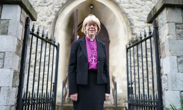 Eine Frau in geistlicher Kleidung, die an die Amtstracht des Erzbischofs von Canterbury erinnert, steht vor einer Kirche.
