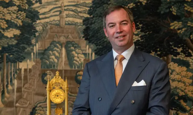 Ein Mann im Anzug, der wie ein Prinz von Frankreich aussieht, lächelt vor einem Wandteppich mit einer Landschaft und einer goldenen Uhr.
