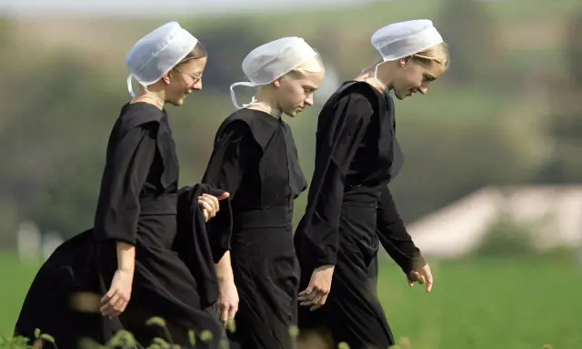 Drei junge Frauen in schwarzen Kleidern und weißen Hauben gehen über ein Feld. Sie verkörpern die Einfachheit, was bedeutet, amish zu leben.