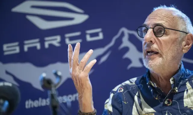 Philip Nitschke spricht mit erhobener Hand vor einem blauen Hintergrund mit dem SARCO-Logo.