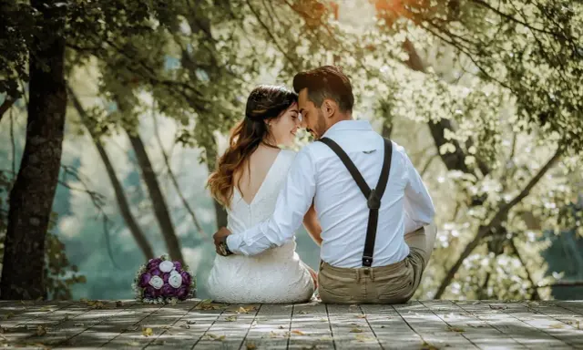 Ein Brautpaar sitzt im Freien, umgeben von Bäumen. Sie halten sich an den Händen und blicken sich liebevoll an. Ein Brautstrauß liegt daneben. Ein Bild, das ihr katholisches Eheversprechen symbolisiert.