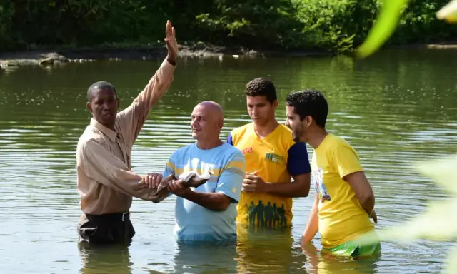 Ein Geistlicher führt eine Baptisten Taufe in einem Fluss durch. Drei Männer stehen im Wasser, während der Geistliche mit erhobener Hand segnet.