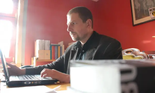 Mann arbeitet am Laptop im Büro des Antaios Verlags. Bücherregale im Hintergrund.