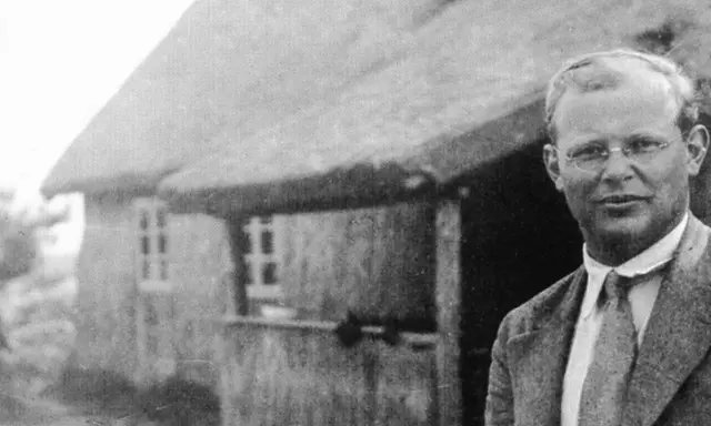 Ein Mann mit Brille steht vor einem Reetdachhaus. Er ist Dietrich Bonhoeffer, ein deutscher Theologe und Widerstandskämpfer.
