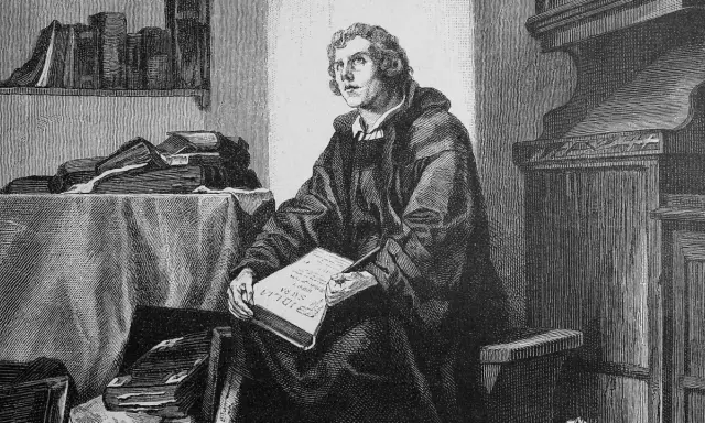 Martin Luther sitzt in seinem Studierzimmer und schreibt an seiner Bibelübersetzung. Bücher stapeln sich um ihn herum.
