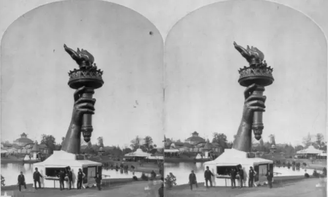 Die Hand mit der Fackel der Freiheitsstatue, ein Werk der freiheitsstatue erbauer, ragt über einem Pavillon auf. Im Hintergrund ist eine Parklandschaft mit einem See und Gebäuden zu sehen.