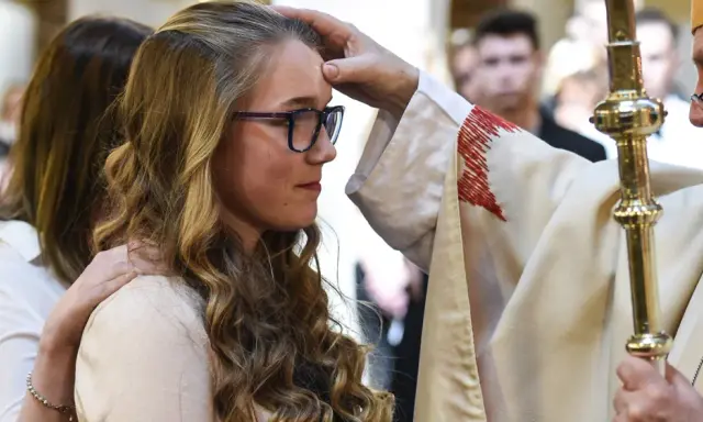 Ein Bischof legt einer jungen Frau die Hand auf die Stirn. Sie hat lange, blonde Haare und trägt eine Brille. Dies ist ein Moment, in dem sie ihren Glauben an Gott bekräftigt.