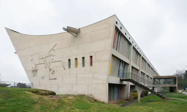 Betonbau mit bunten Fenstern und einer markanten, geschwungenen Form. Ein Meisterwerk des Architekten Le Corbusier.