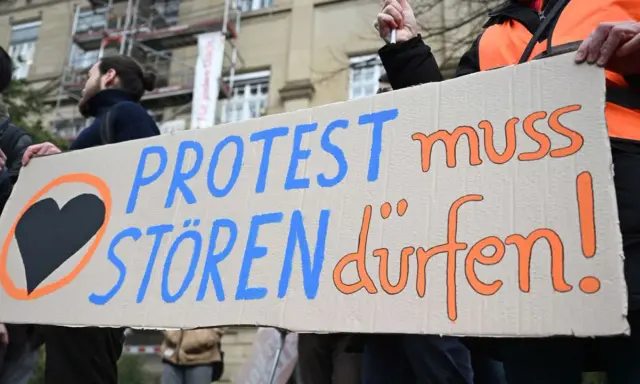Menschen halten ein Schild mit der Aufschrift "PROTEST muss STÖREN dürfen!". Ein schwarzes Herz in einem orangen Kreis symbolisiert ziviler ungehorsam Beispiele.