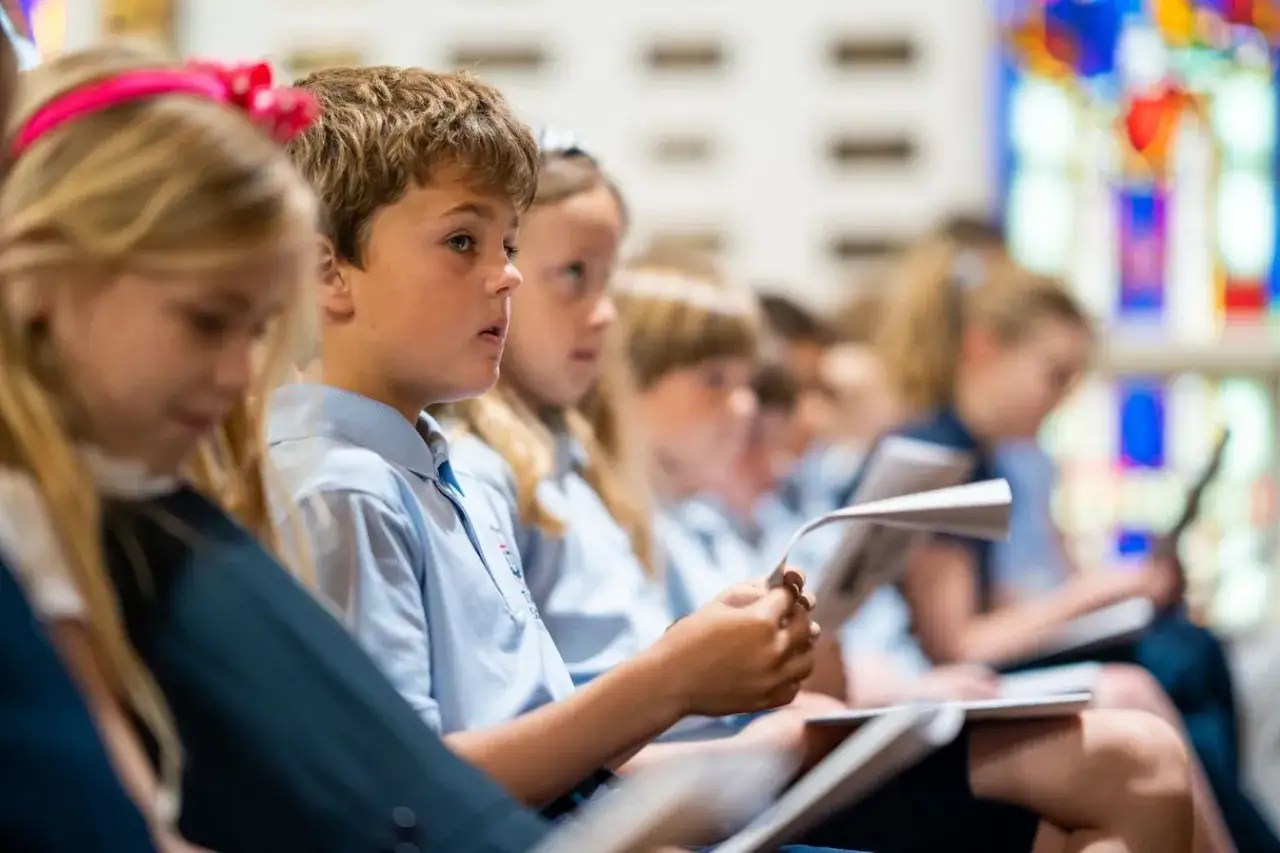 Kinder in Schuluniformen, die in einer Kirche sitzen und singen. Ein Junge im Vordergrund hält ein Notenblatt. Dies ist ein Beispiel für episkopale Erziehung.