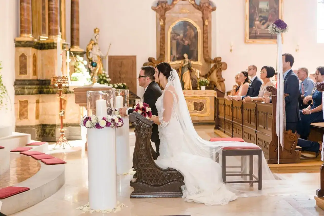 Brautpaar bei katholischer Trauung in einer Kirche, umgeben von Gästen und festlicher Dekoration.