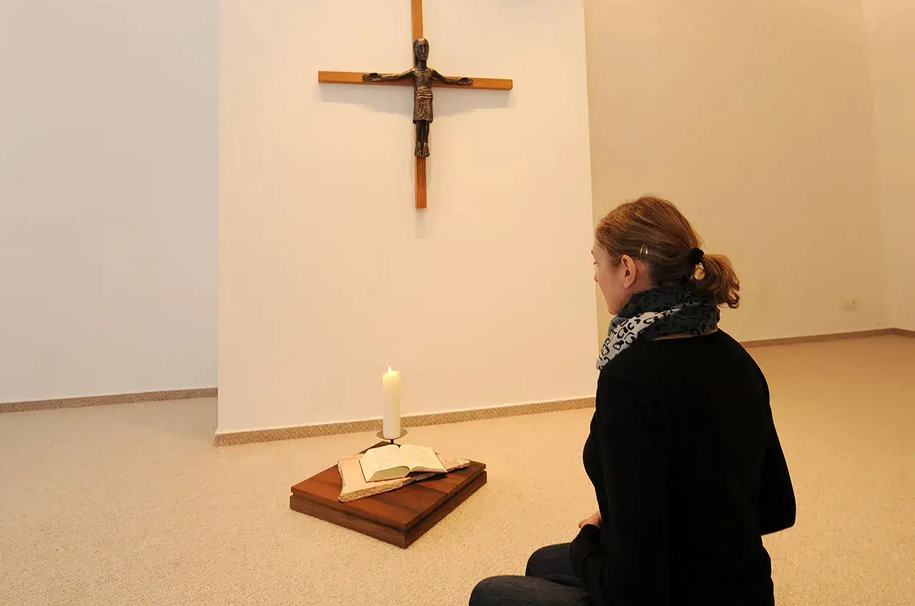 Frau kniet vor einem Kreuz, Kerze und Bibel. Sie sucht Trost und möchte an Gott glauben.