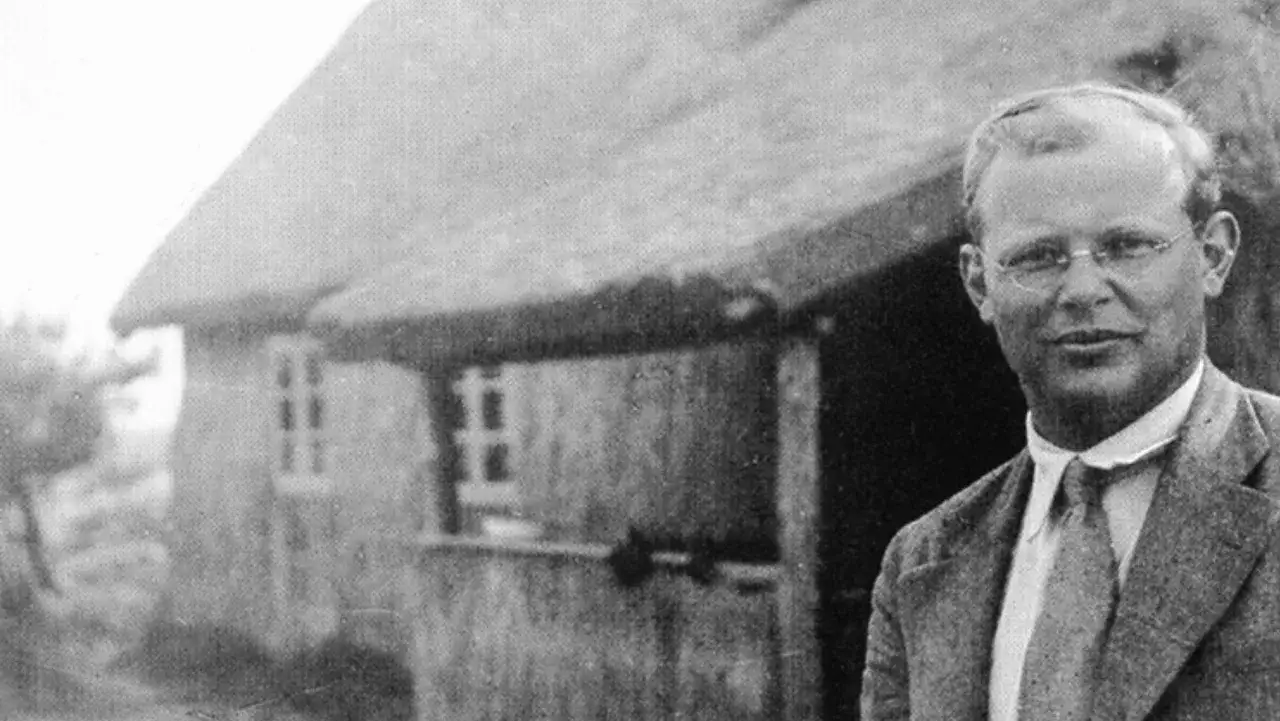 Ein Mann mit Brille steht vor einem Reetdachhaus. Er ist Dietrich Bonhoeffer, ein deutscher Theologe und Widerstandskämpfer.