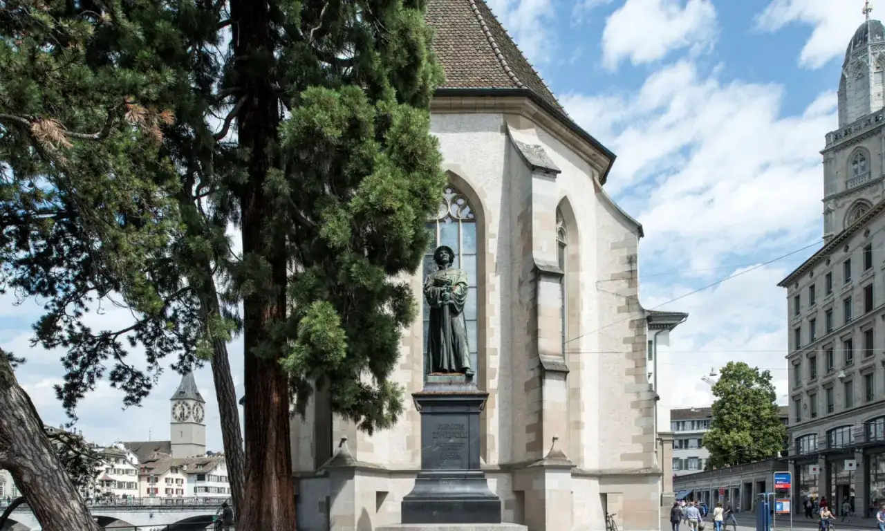 Denkmal für Huldrych Zwingli vor der Kirche, mit Bäumen und Stadtansicht im Hintergrund.