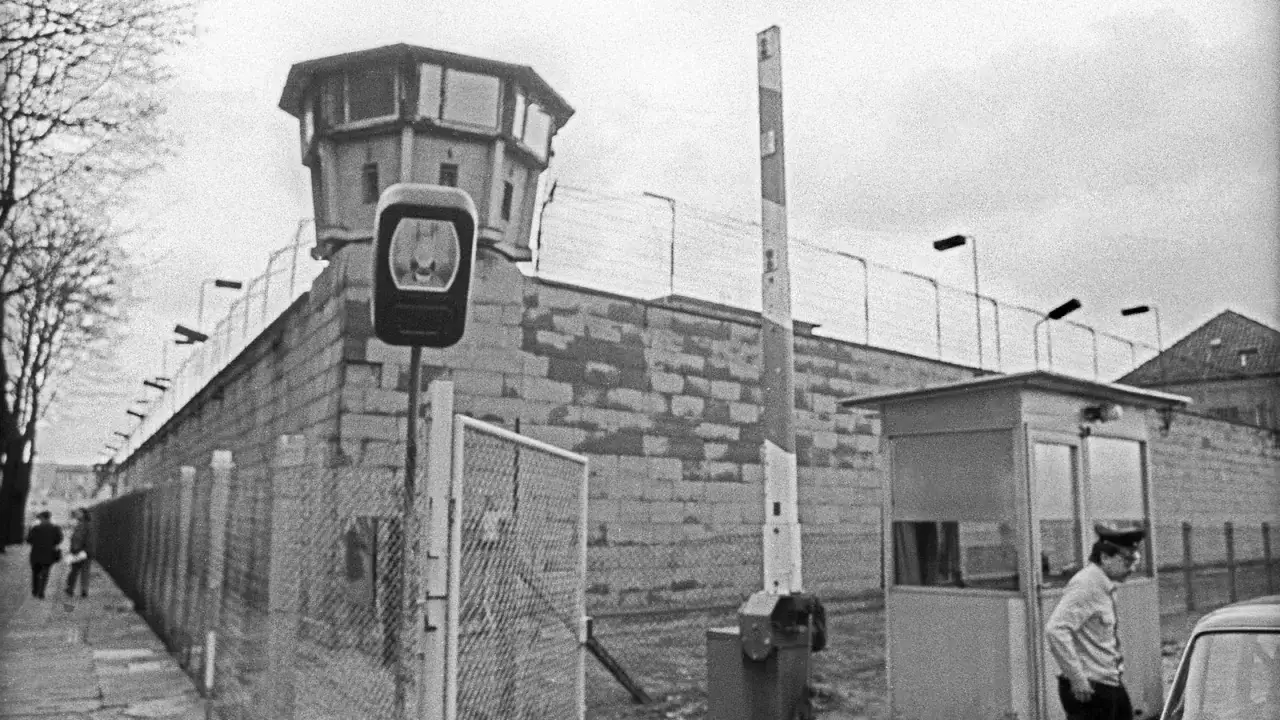 Wachturm und Grenzmauer mit Stacheldraht. Ein Wachmann steht an einer Kontrollbude. Die Szene wirft die Frage auf: war die DDR eine Diktatur?