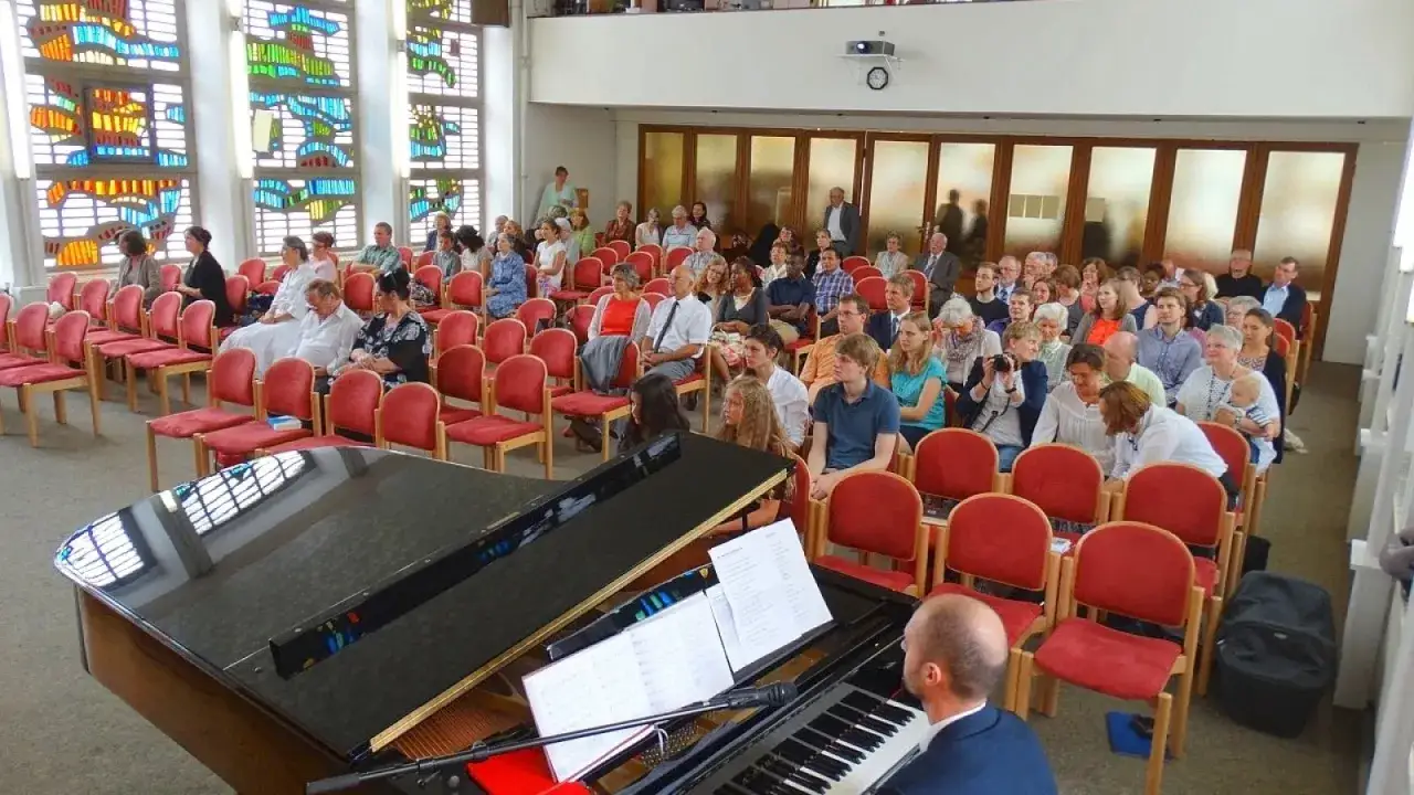 Gemeinde der Siebenten-Tags-Adventisten versammelt sich in einem hellen Kirchenraum mit bunten Glasfenstern und einem Flügel.