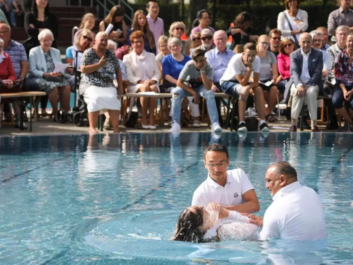 Feierliche Baptisten Taufe im Freien. Ein Geistlicher und ein Helfer tauchen eine Frau in einem weißen Kleid im Pool. Zuschauer sitzen am Beckenrand.