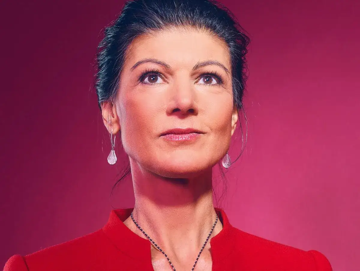 Sahra Wagenknecht, deren Herkunft oft diskutiert wird, blickt nachdenklich nach oben. Sie trägt einen roten Blazer und auffälligen Schmuck.
