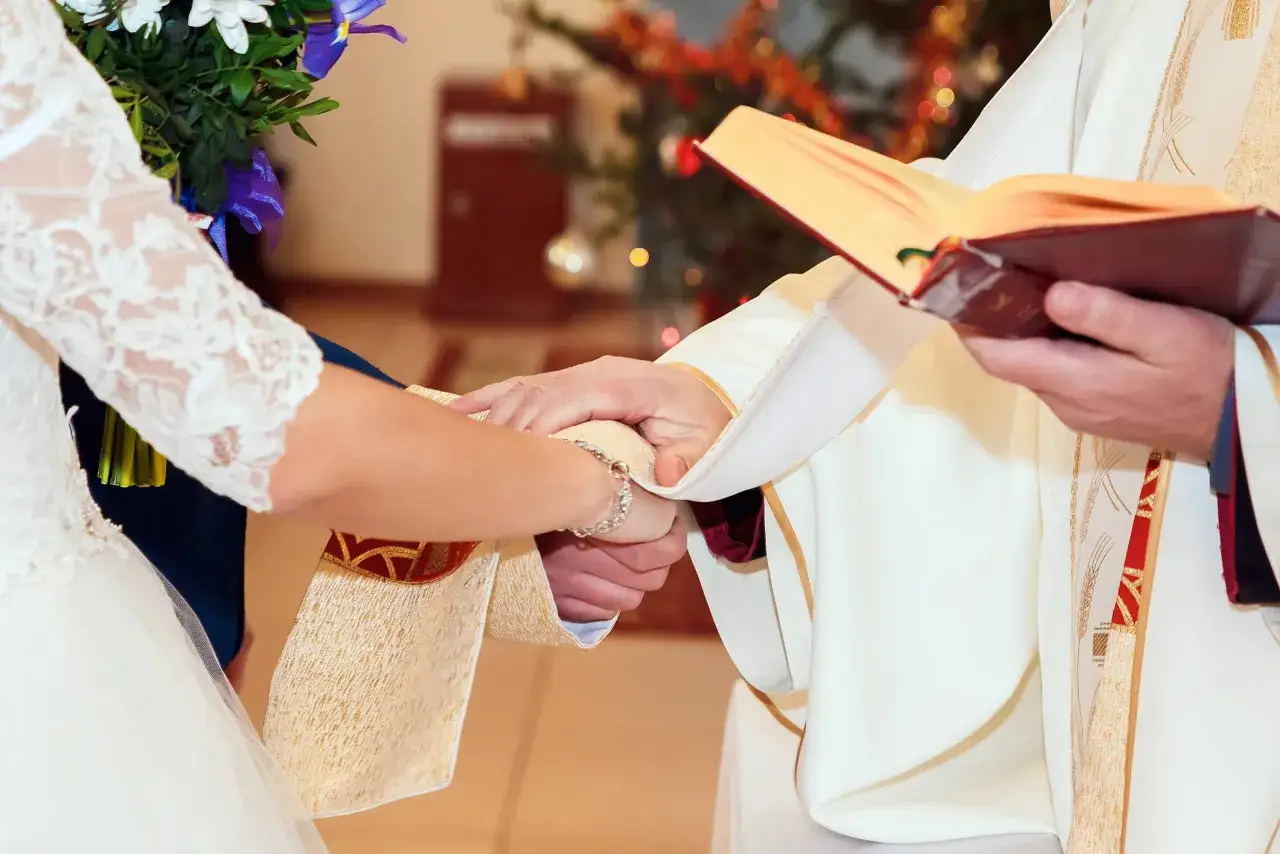 Hände reichen sich bei einem katholischen Eheversprechen. Ein Priester hält ein Buch, während das Brautpaar sich im Hintergrund ein Versprechen gibt.