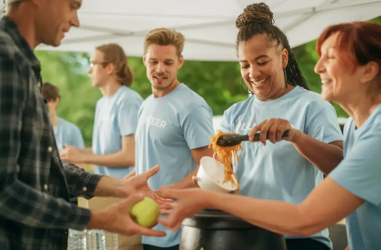 Freiwillige in hellblauen T-Shirts verteilen Essen. Eine Frau lächelt, während sie Nudeln serviert. Dies ist ein Beispiel für altruismus definition in Aktion.