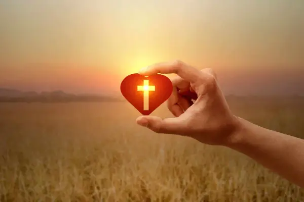Hand hält Herz mit Kreuz, Symbol für christliche Werte im warmen Abendlicht über goldenem Feld.